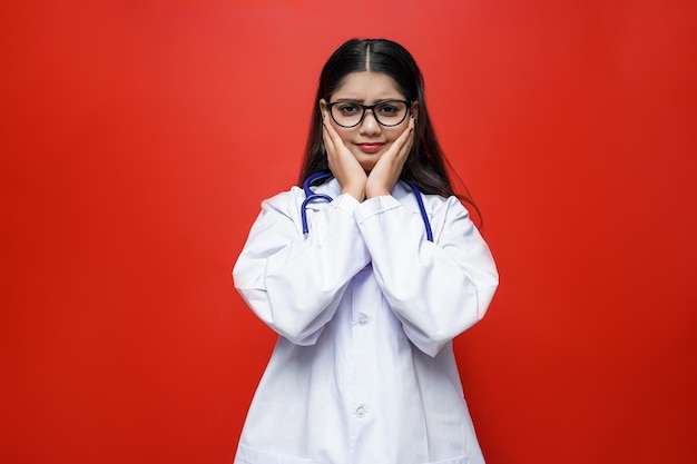 Joven doctora con las manos en las mejillas con gafas sobre fondo rojo modelo paquistaní indio