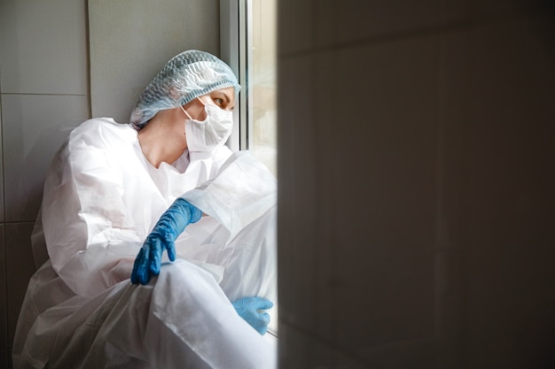 Foto una joven doctora con una máscara de gorra de traje protector y guantes está sentada con cansancio cerca de la ventana en ...