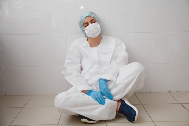Una joven doctora en una máscara de gorro de traje protector y guantes está sentada con cansancio en el suelo