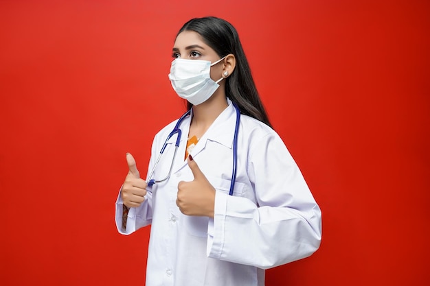 Joven doctora con máscara muestra Thumbs-up sobre fondo rojo modelo paquistaní indio