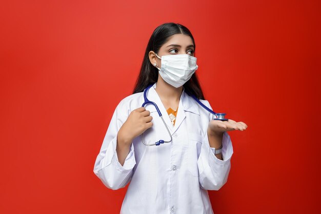Joven doctora con máscara, sosteniendo un estetoscopio sobre fondo rojo modelo paquistaní indio