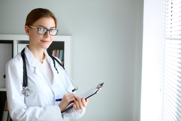 Joven doctora morena de pie con portapapeles cerca de la ventana en el hospital. Médico listo para examinar al paciente