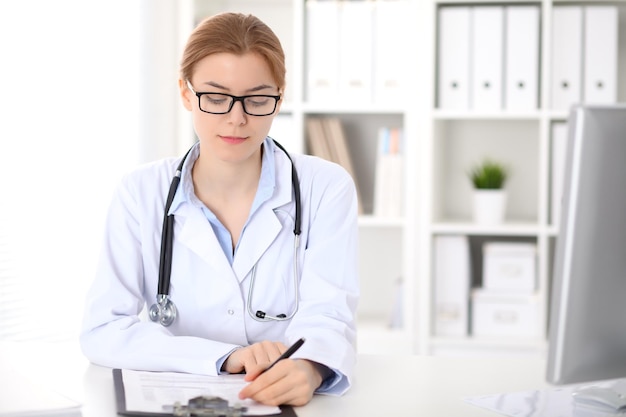 Joven doctora morena sentada en la mesa y trabajando en la oficina del hospital. Concepto de atención médica, seguro y ayuda. Médico listo para examinar al paciente