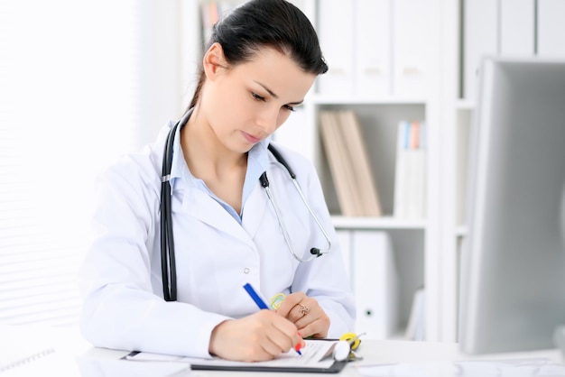 Joven doctora morena sentada con portapapeles cerca de la ventana en el hospital y llenando el formulario de historial médico.