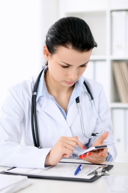 Joven doctora morena sentada con portapapeles cerca de la ventana en el hospital y llenando el formulario de historial médico.