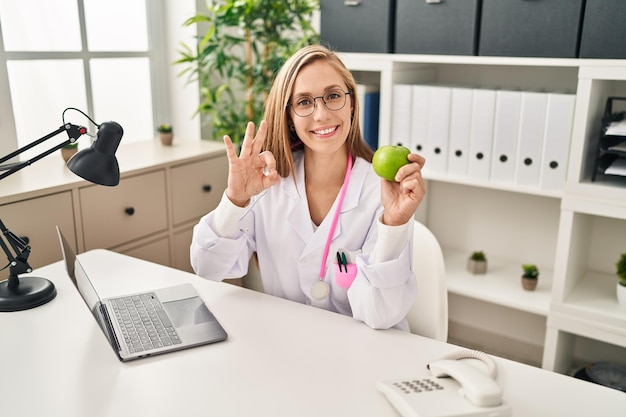 Joven doctora rubia sosteniendo manzana verde en la clínica haciendo el signo de ok con los dedos sonriendo amigablemente gesticulando símbolo excelente