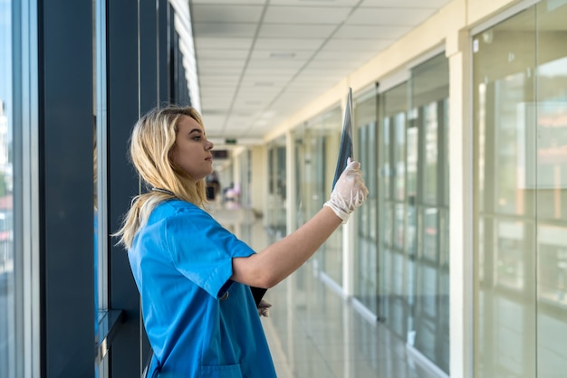 Joven doctora rubia vestida con uniforme azul con estetoscopio sosteniendo una película de radiografía de tórax y controlando la neumonía. COVID-19