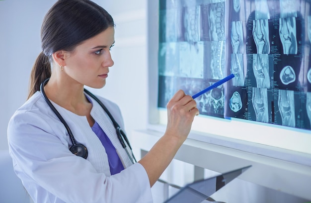 Joven doctora sonriente con estetoscopio apuntando a rayos X en el consultorio del médico