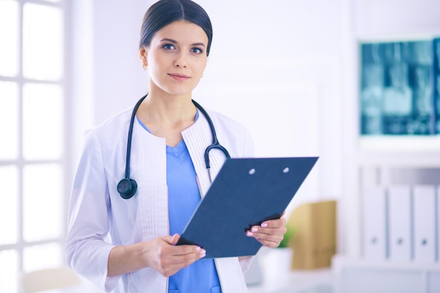 Joven doctora sonriente con estetoscopio sosteniendo una carpeta en el consultorio del médico