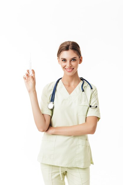 Joven doctora sonriente en uniforme con fonendoscopio en el cuello sosteniendo la jeringa en la mano mientras soñaba