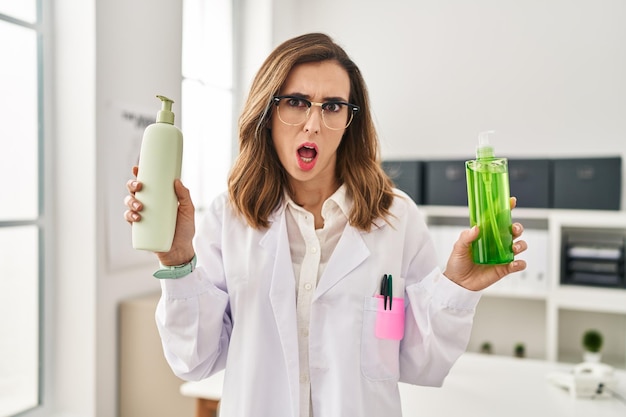 Joven doctora sosteniendo aloe vera y crema en la cara de shock, luciendo escéptica y sarcástica, sorprendida con la boca abierta