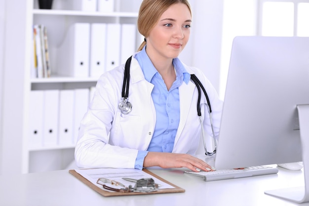 Joven doctora en el trabajo en el hospital mirando el monitor de pc de escritorio. El médico controla los registros del historial de medicamentos y los resultados de los exámenes. Concepto de medicina y salud.