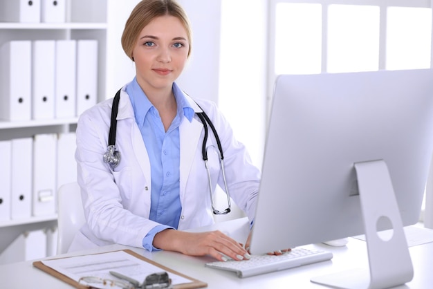 Joven doctora en el trabajo en el hospital mirando el monitor de pc de escritorio. El médico controla los registros del historial de medicamentos y los resultados de los exámenes. Concepto de medicina y salud.