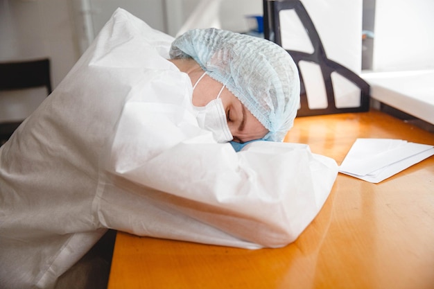 Foto una joven doctora con un traje protector, máscara, gorro y guantes yace cansada en la mesa con sus ojos ...