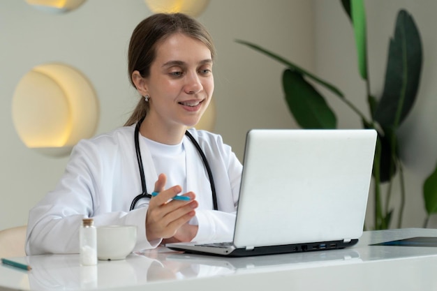 Una joven doctora en un uniforme médico blanco con un estetoscopio usando una computadora portátil está hablando a través de