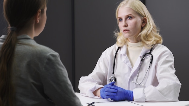 Joven doctora en uniforme médico blanco que usa el portapapeles está brindando una gran charla de noticias sobre los resultados o los síntomas con una paciente