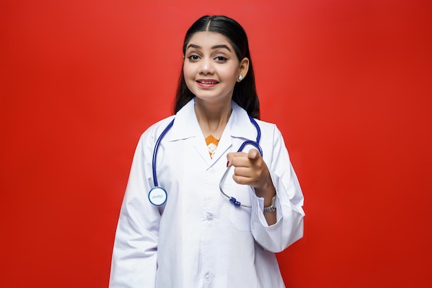 Joven doctora vistiendo abrigo y estetoscopio apuntando hacia el frente con una sonrisa modelo paquistaní indio