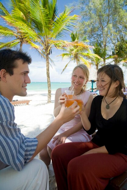 Un joven y dos mujeres jóvenes diciendo saludos con sus cócteles en la playa