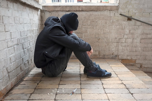 Un joven drogadicto en un capó se sienta en una casa abandonada después de tomar el concepto de adicción a las drogas