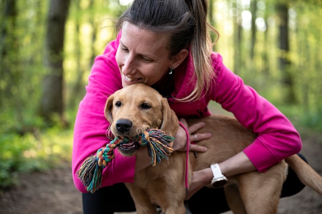 Joven dueña de perro en suéter rosa acurrucada con su cachorro de labrador retriever de pura raza en el bosque de primavera verde