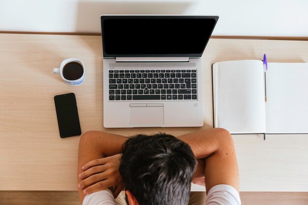 Foto joven durmiendo en la mesa frente a la computadora apoyado en sus brazos con un café, un teléfono inteligente y un cuaderno concepto de teletrabajo o clases en línea