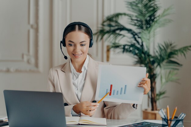 Joven economista empleada en traje elegante y auriculares con presentación en línea, apuntando al informe financiero con estadísticas en aumento durante la videollamada en la computadora portátil y sintiéndose satisfecha con los resultados