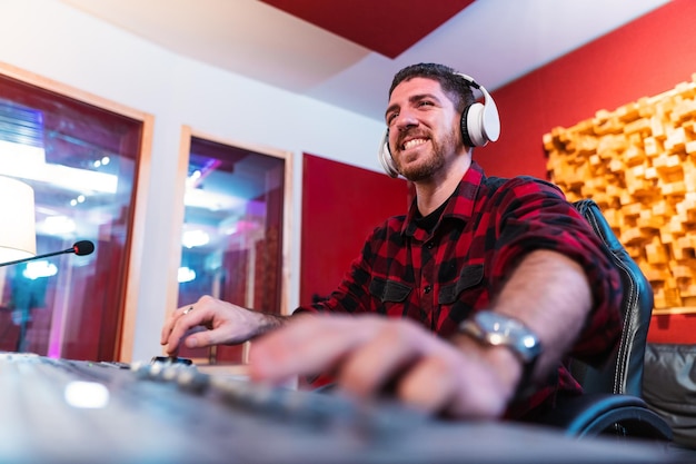 Joven ecualizando la canción en un estudio de música profesional usando una computadora usando auriculares Feliz ingeniero de sonido trabajando y mezclando pista Concepto de tecnología