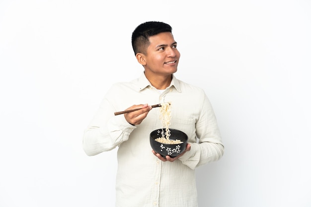 Joven ecuatoriano aislado sobre fondo blanco sosteniendo un plato de fideos con palillos y mirando hacia arriba