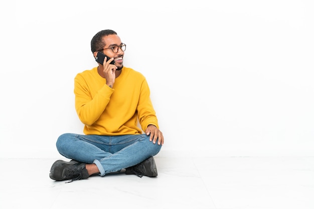 Joven ecuatoriano sentado en el suelo aislado en una pared blanca manteniendo una conversación con el teléfono móvil con alguien