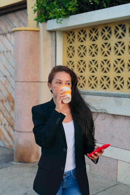 Joven ejecutiva tomando café y sosteniendo su teléfono celular mientras está de pie en el tiempo libre