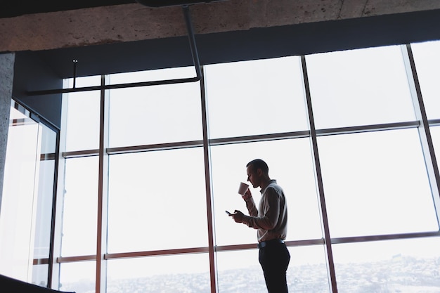 Un joven ejecutivo sostiene un teléfono mientras está parado en el interior de una oficina y mira por una ventana grande con vista a la ciudad El gerente masculino con un teléfono está pensando en un nuevo proyecto de negocios