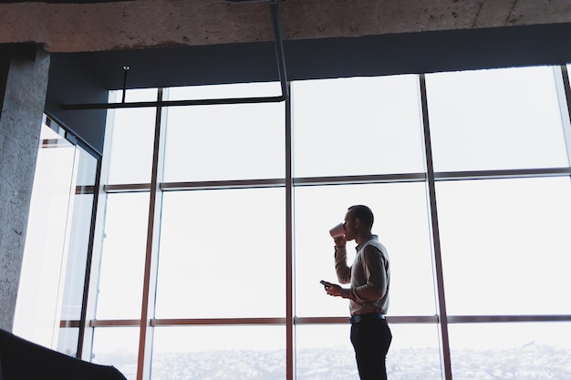 Un joven ejecutivo sostiene un teléfono mientras está parado en el interior de una oficina y mira por una ventana grande con vista a la ciudad El gerente masculino con un teléfono está pensando en un nuevo proyecto de negocios