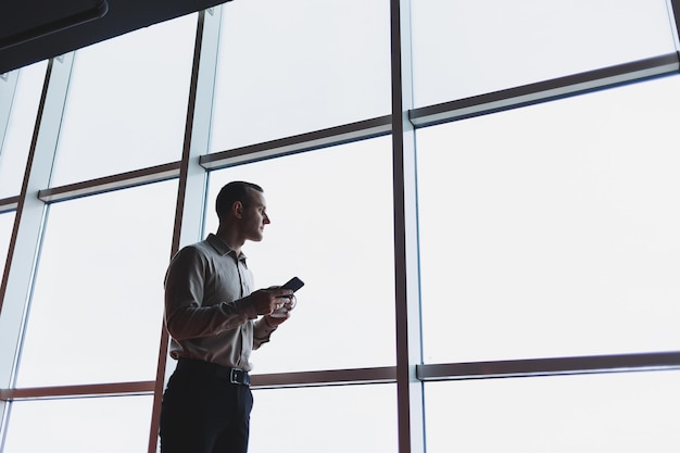 Un joven ejecutivo sostiene un teléfono mientras está parado en el interior de una oficina y mira por una ventana grande con vista a la ciudad El gerente masculino con un teléfono está pensando en un nuevo proyecto de negocios