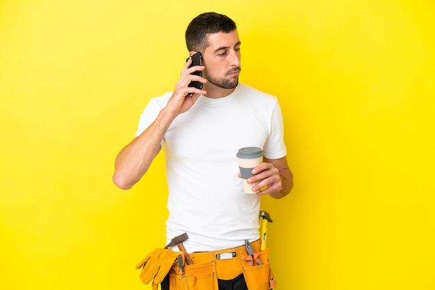 Joven electricista caucásico aislado de fondo amarillo sosteniendo café para llevar y un móvil