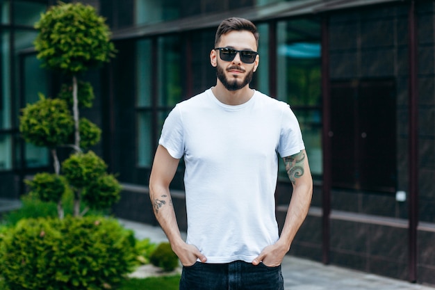 Un joven elegante con barba en una camiseta blanca y gafas de sol