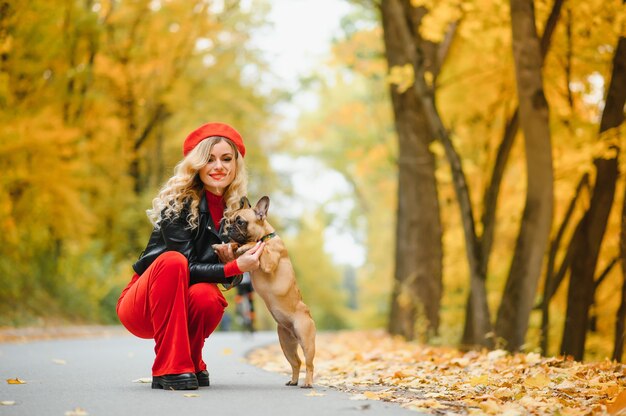 Una joven elegante con cabello largo y claro con gafas soleadas sale a caminar con un perrito mediano, un pug junto al bulldog francés en un parque en primavera en otoño