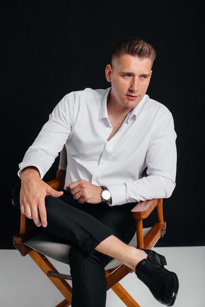 Un joven elegante con una camisa blanca está sentado en la silla del director sobre un fondo negro. Un apuesto hombre de negocios carismático.