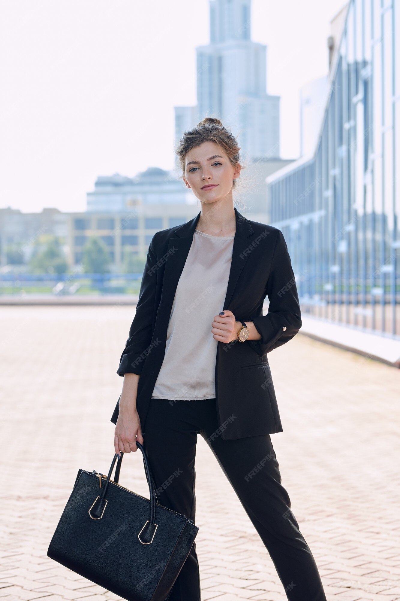 Joven y elegante dama de negocios rubia en ropa casual elegante sosteniendo  un bolso de cuero negro mientras está de pie frente a la cámara contra el  paisaje urbano | Foto Premium