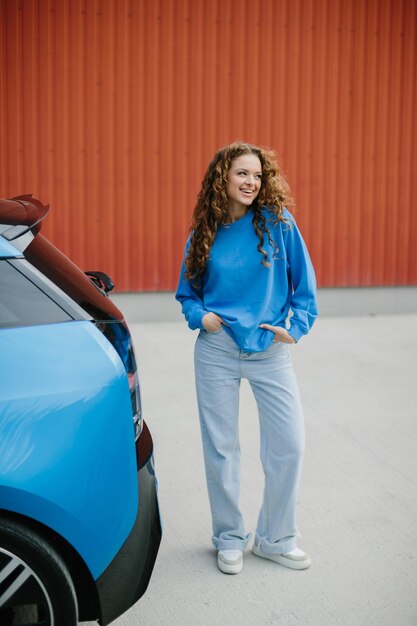 Una joven elegante está de pie al lado de su coche eléctrico en el estacionamiento