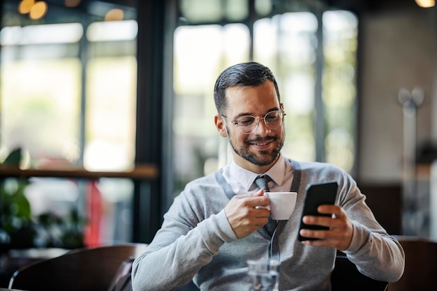 Un joven elegante feliz está tomando café en la cafetería y usando su teléfono