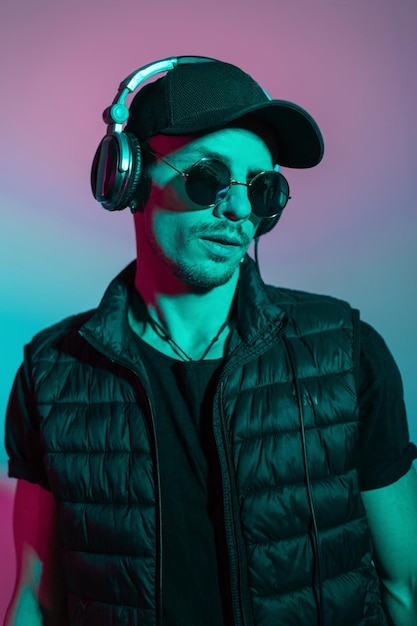 Un joven elegante con gafas de sol de moda y auriculares con ropa exterior negra con gorra y chaleco en el estudio con luz azul y rosa de color El chico escucha música