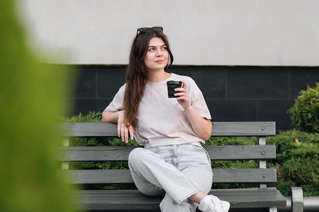 Una joven elegante con gafas de sol se sienta en un banco y bebe café