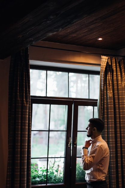 Un joven y elegante novio con traje azul y camisa blanca se para en la habitación y mira por la ventana