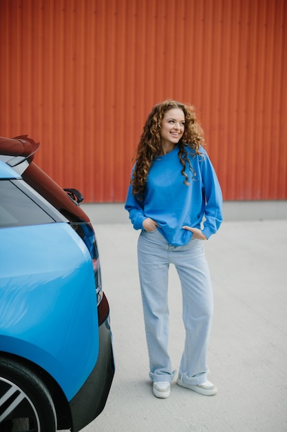 Foto una joven elegante está de pie al lado de su coche eléctrico en el estacionamiento