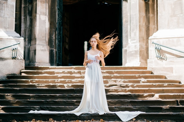 Joven elegante romántica con cabello al viento en vestido largo blanco posando en las escaleras del antiguo palacio
