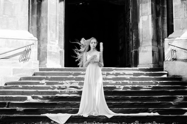 Joven elegante romántica con cabello volador en vestido largo blanco posando en las escaleras del antiguo palacio