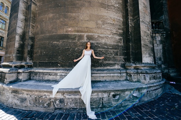 Joven elegante romántica en vestido blanco largo posando sobre muro antiguo de piedra