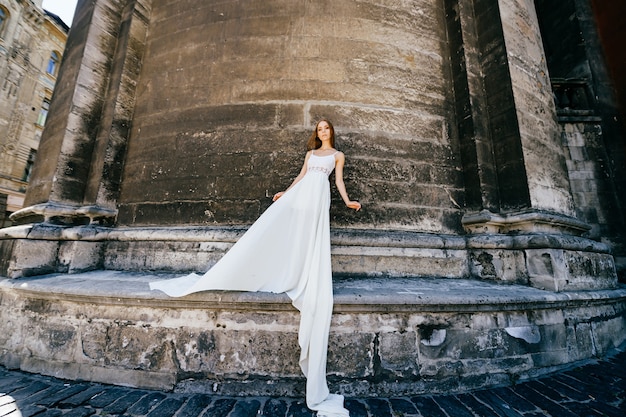 Joven elegante romántica en vestido blanco largo posando sobre muro antiguo de piedra