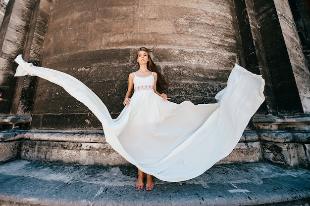 Foto joven elegante romántica en vestido largo blanco fluido posando sobre muro antiguo de piedra