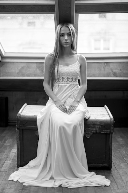 Joven elegante romántica en vestido largo blanco sentado en el pecho en el desván